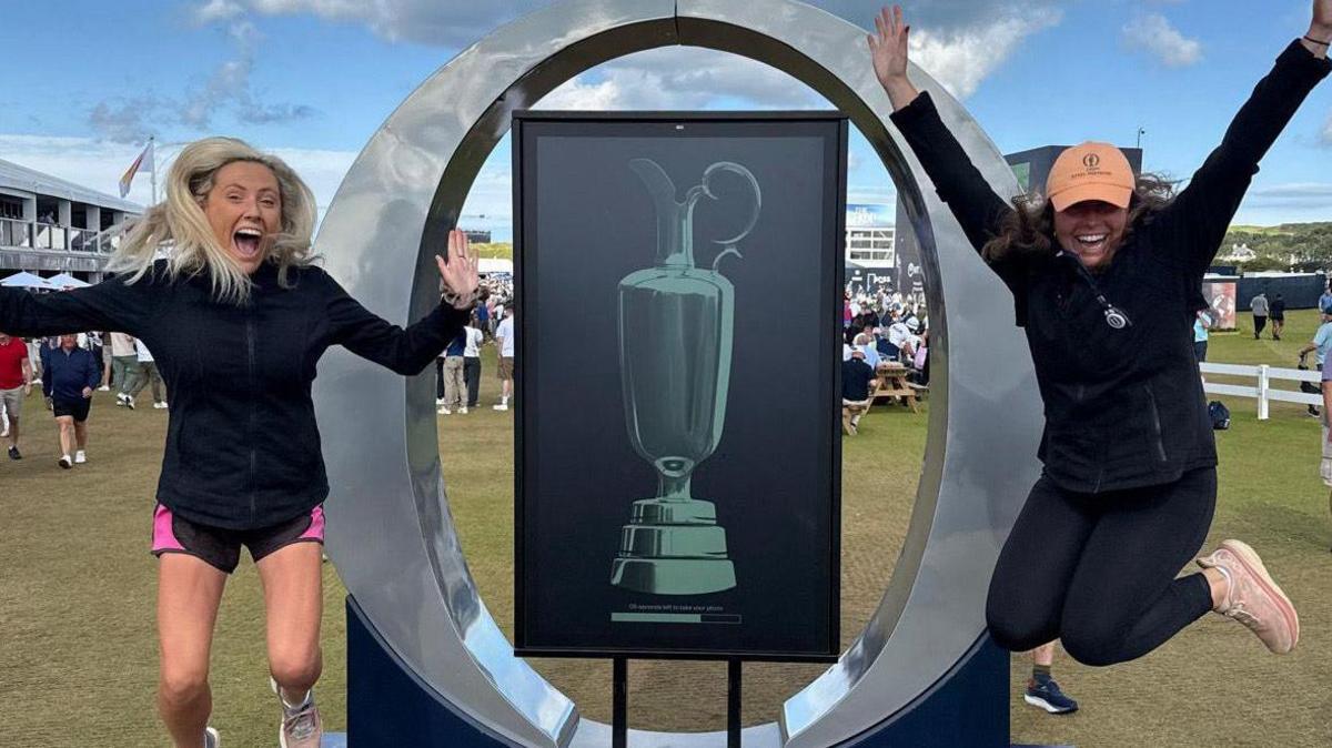 Two smiling woman are seen jumping in the air with their hands raised above their head. One of the woman is wearing pink shorts and a black long sleeve top. The other woman is wearing black leggings and a black long sleeve fleece and an orange coloured base ball cap. They are positioned on either side of a silver statue of the letter O to symbolise The Open golf championship. There is a picture inside the O of the Claret Jug.