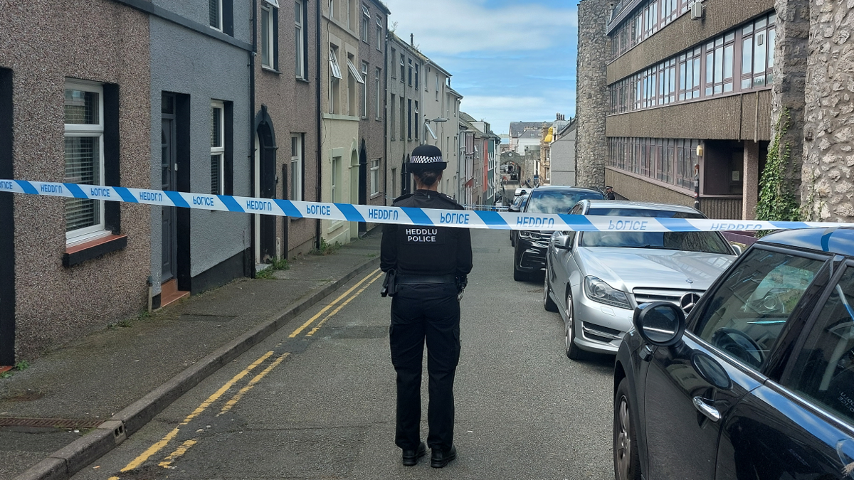 A police officer in a closed terraced street in Caernarfon, near vehicles, and with blue police tape across the road 