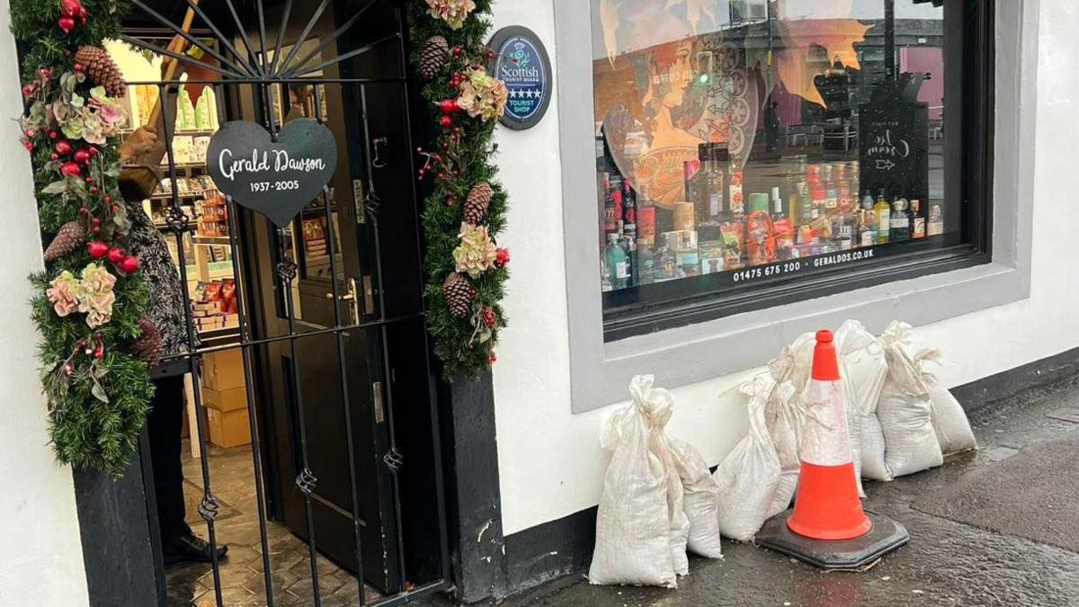 The front of a shop. Sandbags and a traffic cone are placed against the front wall to try and protect it from the rain.