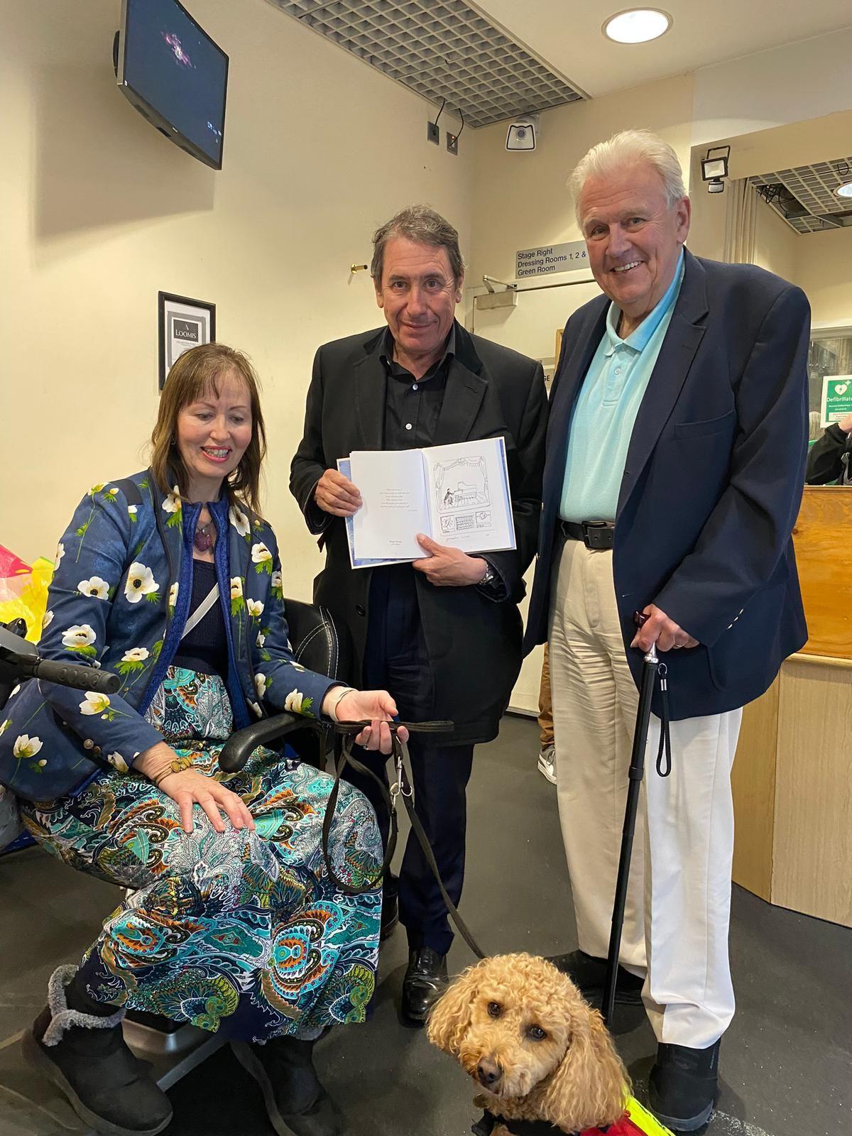 A woman sat in a mobility scooter holding a golden poodle on dog lead. Two men are stood to her right wearing blazers and smiling.