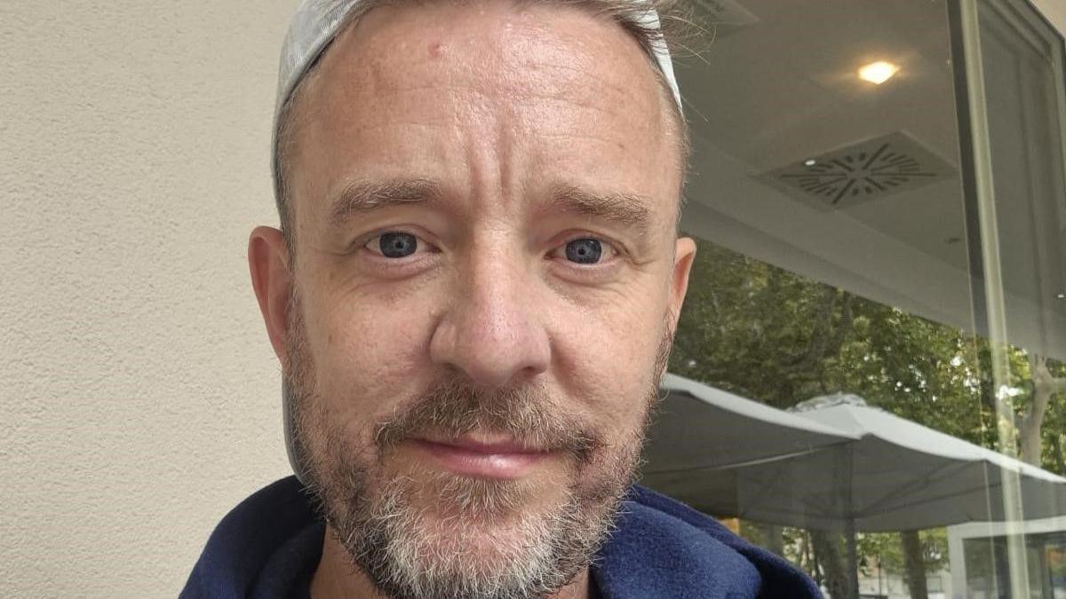 Man looking directly at camera. He has blue eyes and a grey and dark beard. He has fine, short hair, and is wearing a white hat. 