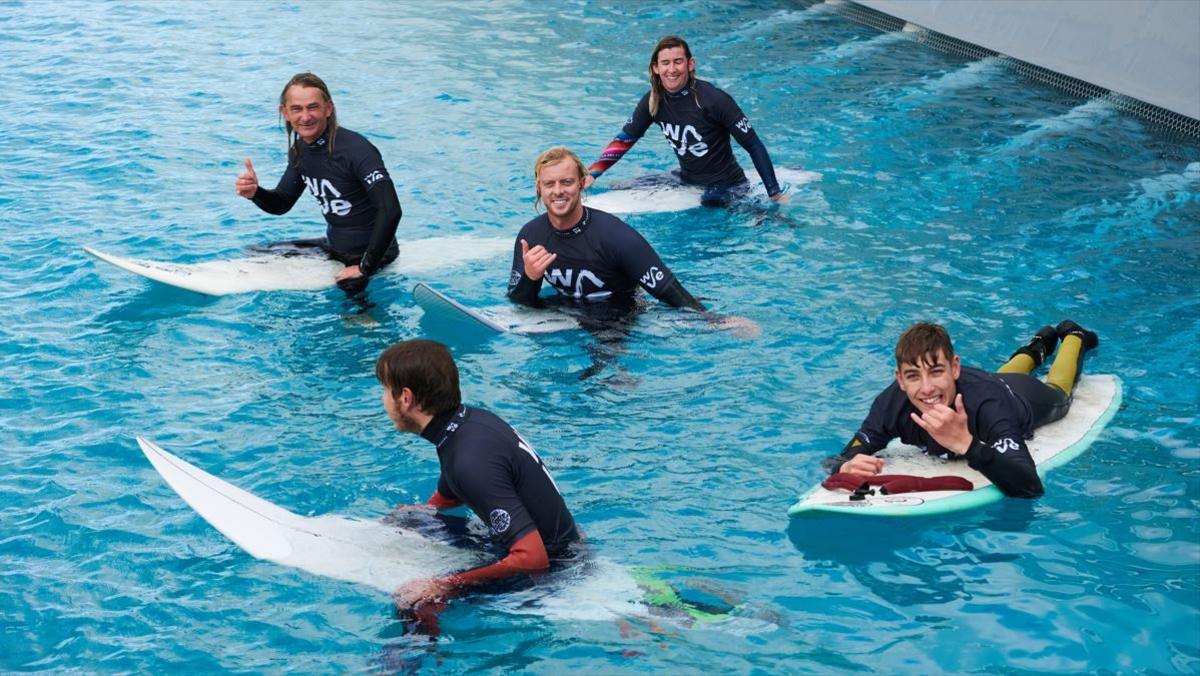 Five male surfers sitting or lying on their white surfboards in the water at The Wave. They are all wearing black wetsuits and black rash vests which say 'The Wave' on them. They all have very wide smiles and a few of them are signing 'shaka', which is a common expression in the surfing industry. 