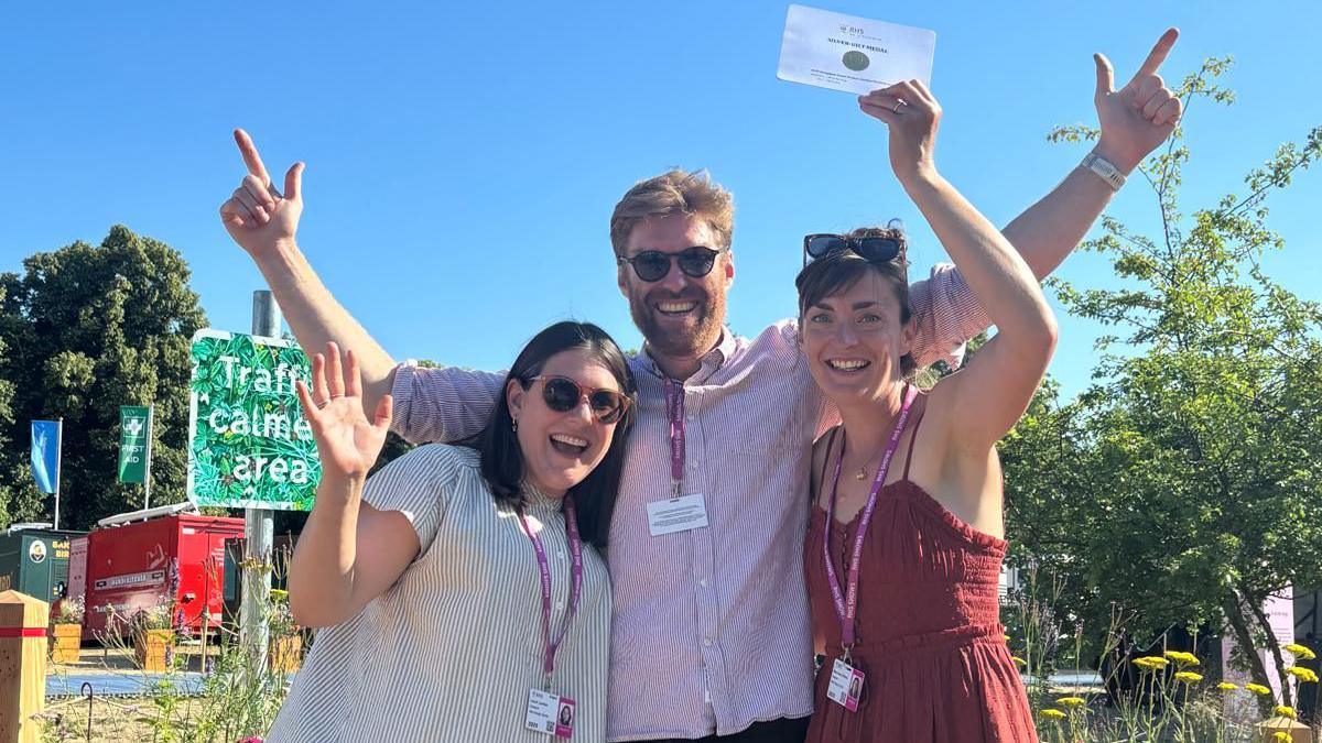 Mary-Anne O'Brien, Robin Dunlop and Laura London stood together in the sun. One of them holds a white certificate in the air and they are all smiling. Behind them are plants that form their garden show exhibition.