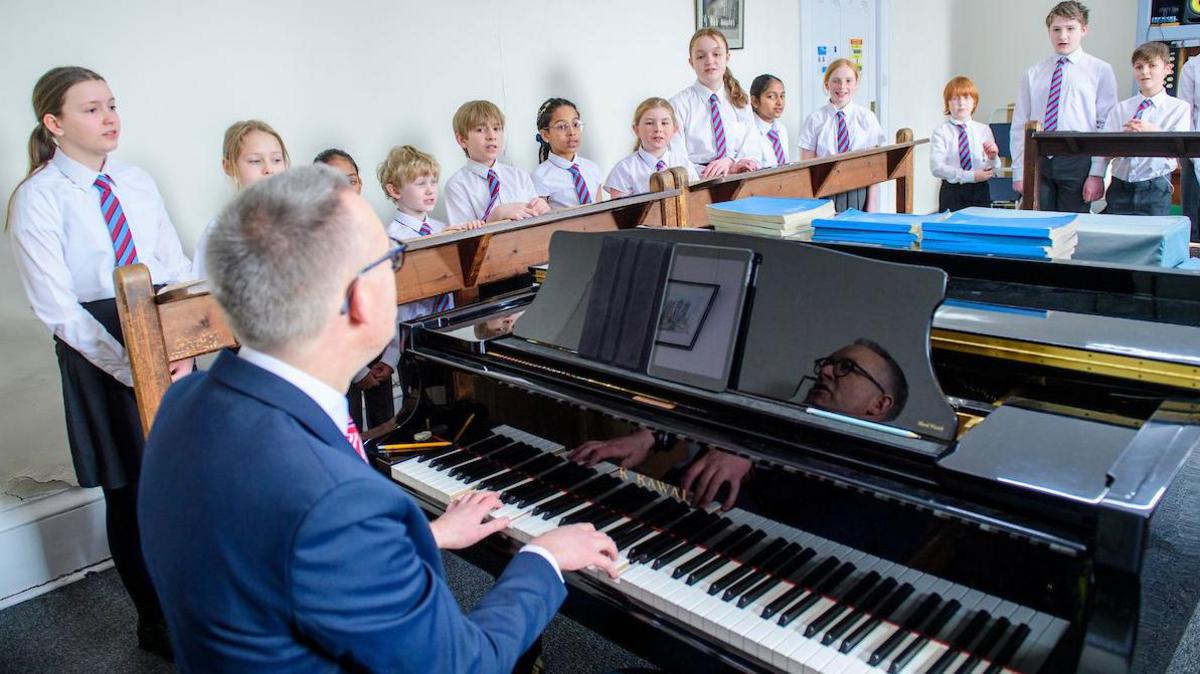 A man in a blue jacket plays the piano to accompany singing by a group of schoolchildren.