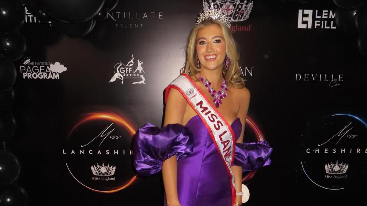 Tabitha stands in front of a black backdrop with small sponsorship logos on it. She wears a purple off the shoulder gown with large, puffy sleeves. She has long blonde hair and wears a silver crown and a red and silver sash. 