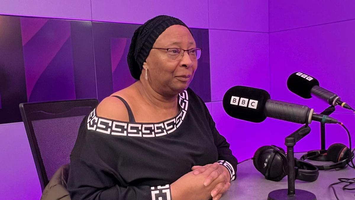 Marsha sits at a desk in the BBC Radio Bristol studio. She wears a long-sleeved black top, black glasses and has a hair covering on her head.