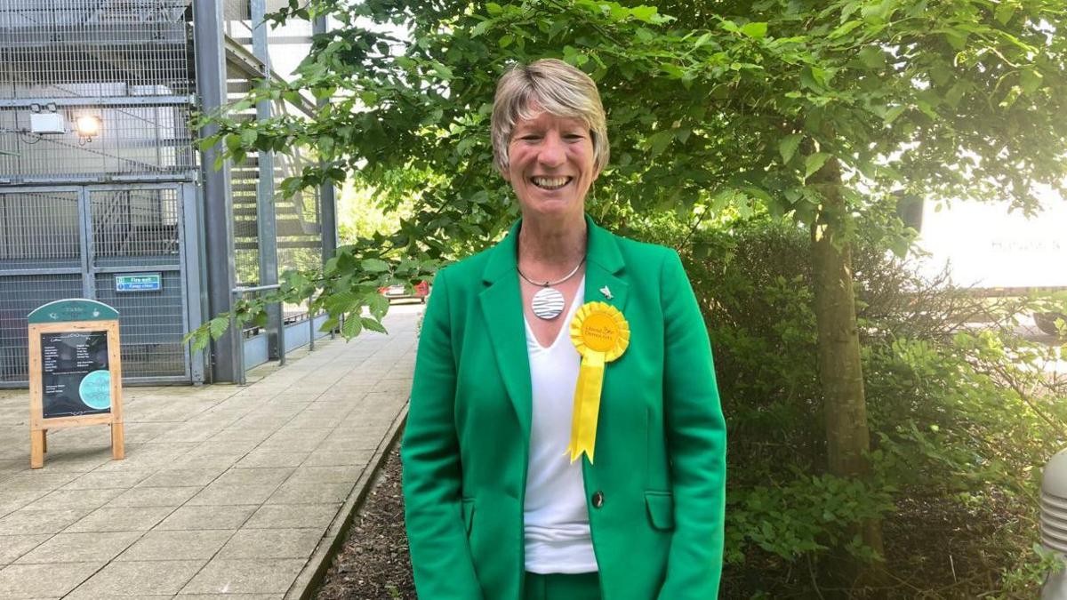Pippa Heylings looks at the camera with a smile on her face. She wears a green jacket, a yellow Liberal Democrat rosette and a necklace with a large circle pendant. In the background is a tree with lots of green leaves. To the left is a sign advertising coffee for sale.