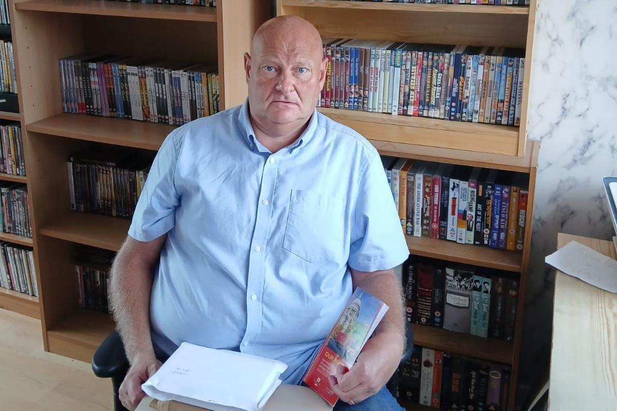 Mark Corfield is sitting in front of a bookcase full of DVDs with a pile of parcels on his lap.
