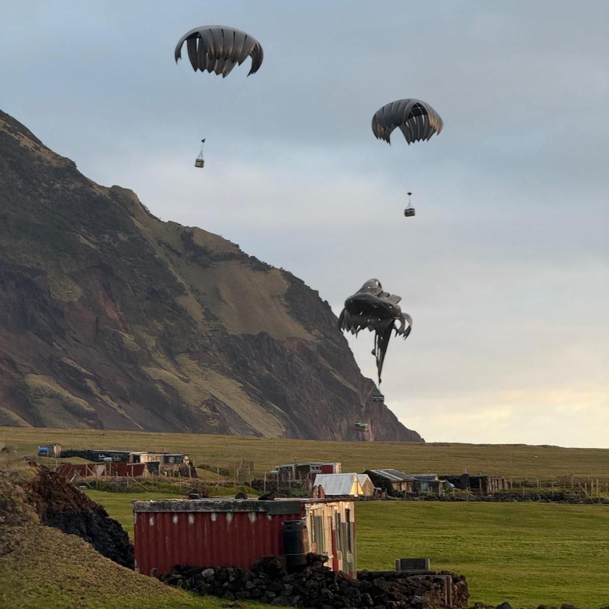 Parachutes delivering boxes of military supplies seen against a rugged backdrop, where several small buildings can be seen