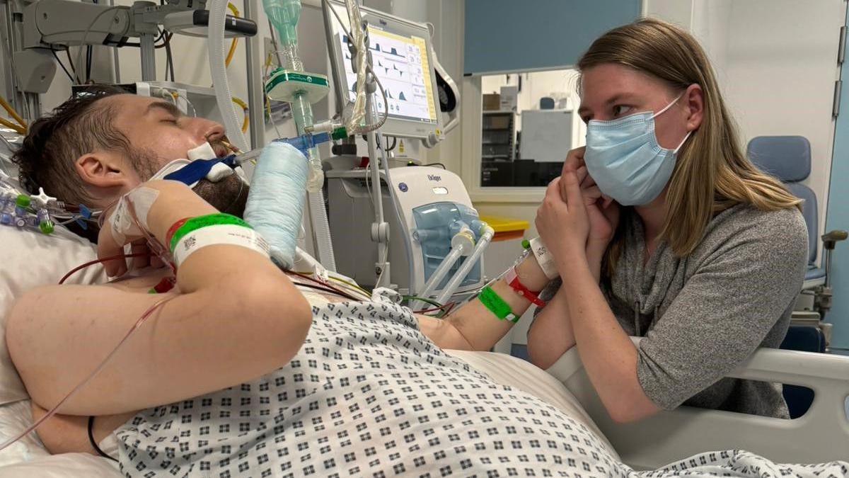 Laura Burr sits by her husband's hospital bed, holding his hand, on the day before he died. She wears a disposable face mask. He is intubated and apparently asleep.