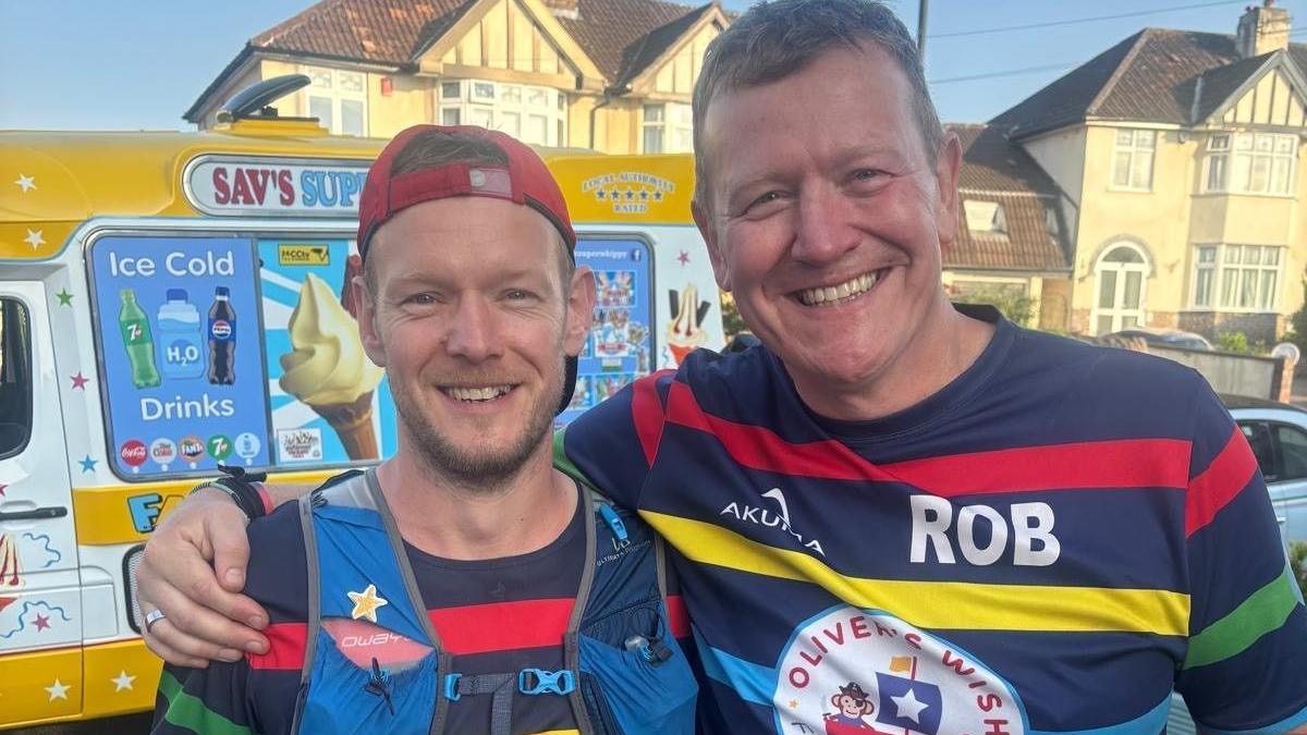 Rob Dowling, pictured on the right, has his hand around Simon Pilkington's shoulder as they smile at the camera, They are wearing their Oliver's Wish t-shirts and stand in front of an ice cream van.