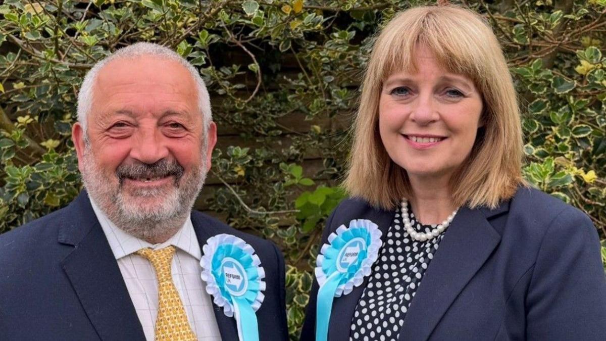 A man and a woman are wearing light blue and white rosettes featuring the Reform UK logo. The man, on the left with grey hair and a beard, and the woman, with blonde/brown hair, are both wearing blue jackets.
