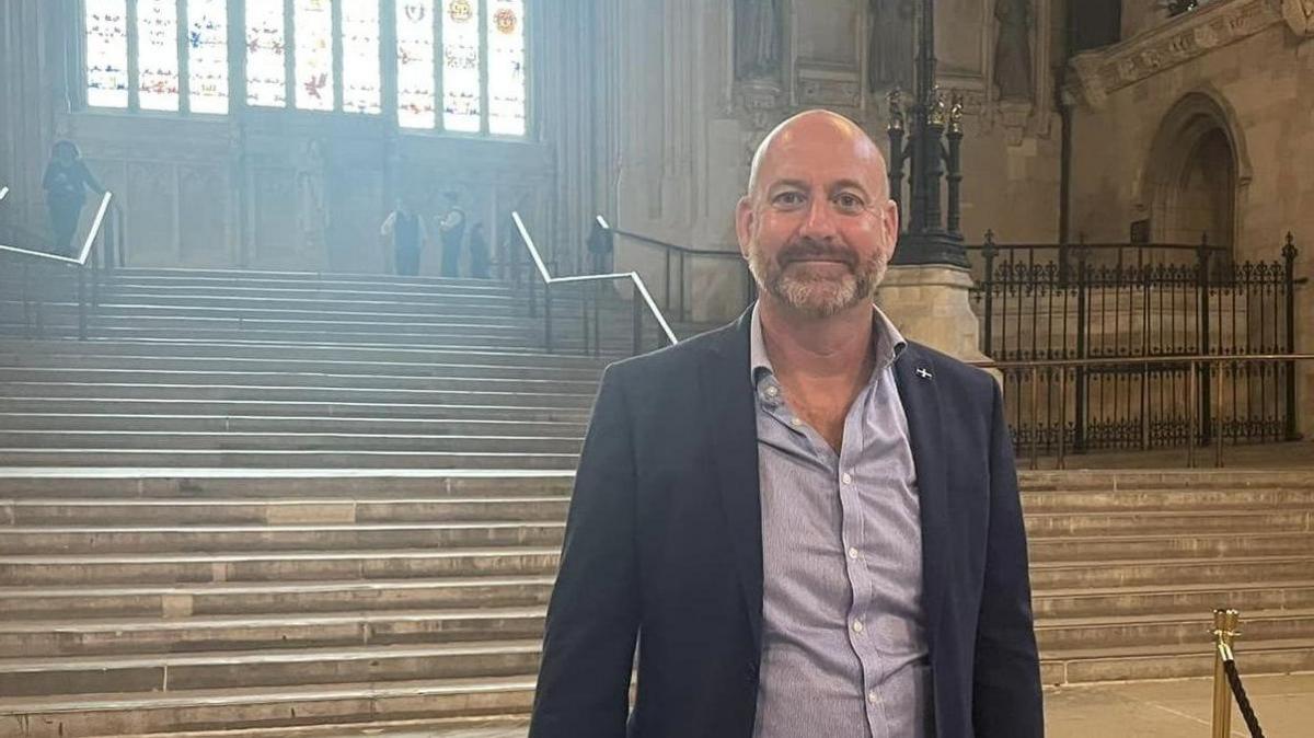 Perran Moon is standing in the Houses of Parliament. He is smiling and wearing a blue shirt and jacket with a St Piran flag buttonhole badge. Behind him are a set of wide stone steps which lead up to a stained glass window. The stone walls are carved and there is an archway behind ornate iron railings. At the top of the steps are two security officers talking to each other.