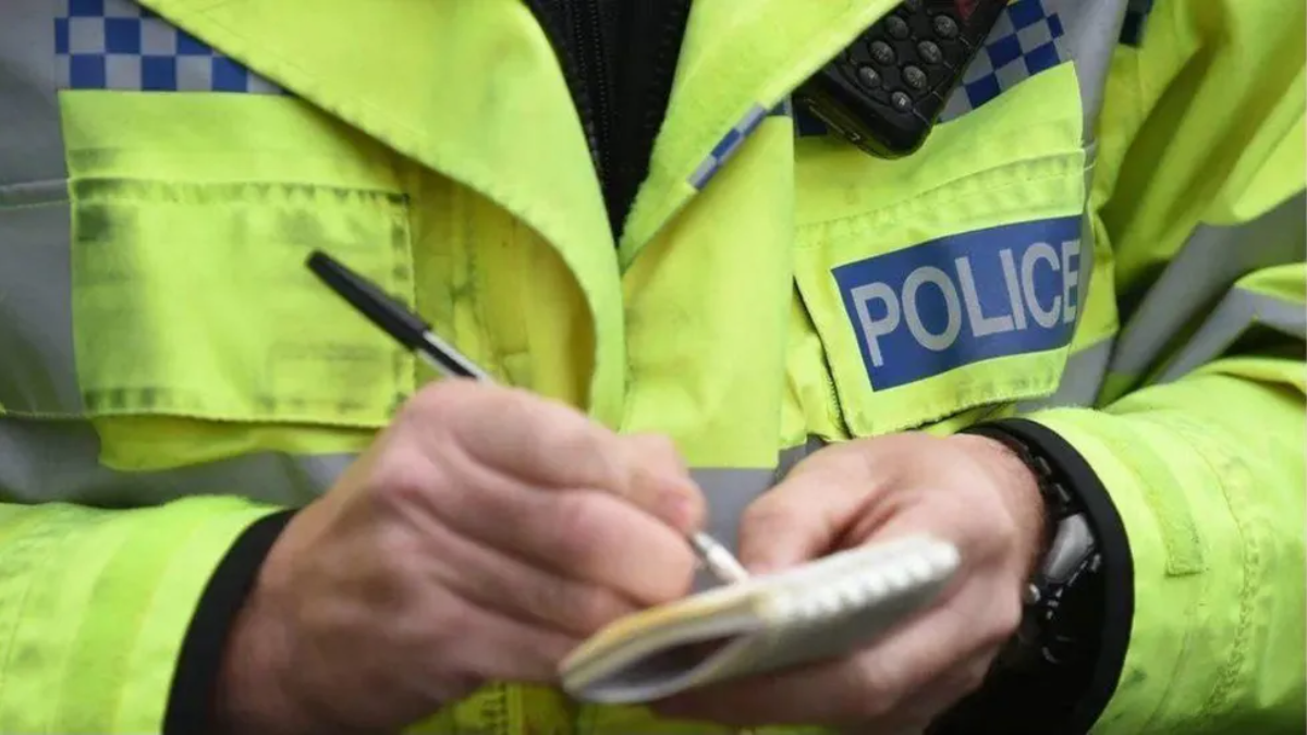 Police officer wearing bright yellow police jacket writing notes. There is a radio walkie-talkie on their chest.