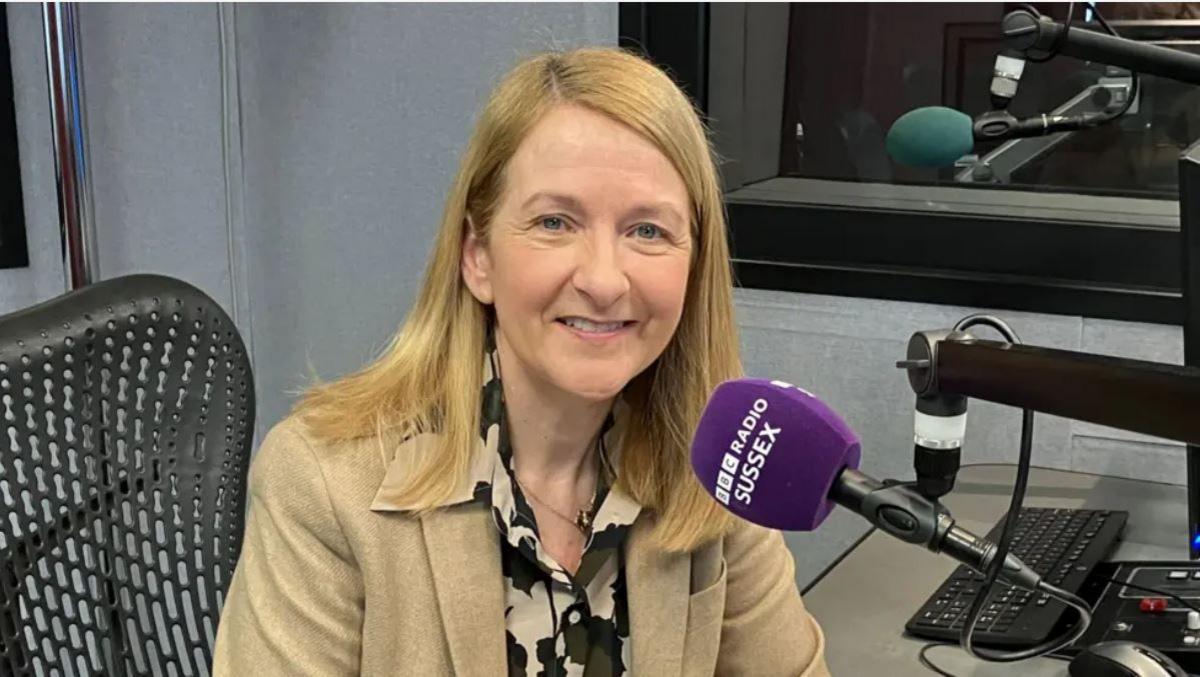 A woman with blonde hair is sitting at a desk and smiling at the camera. She is wearing a beige jacket