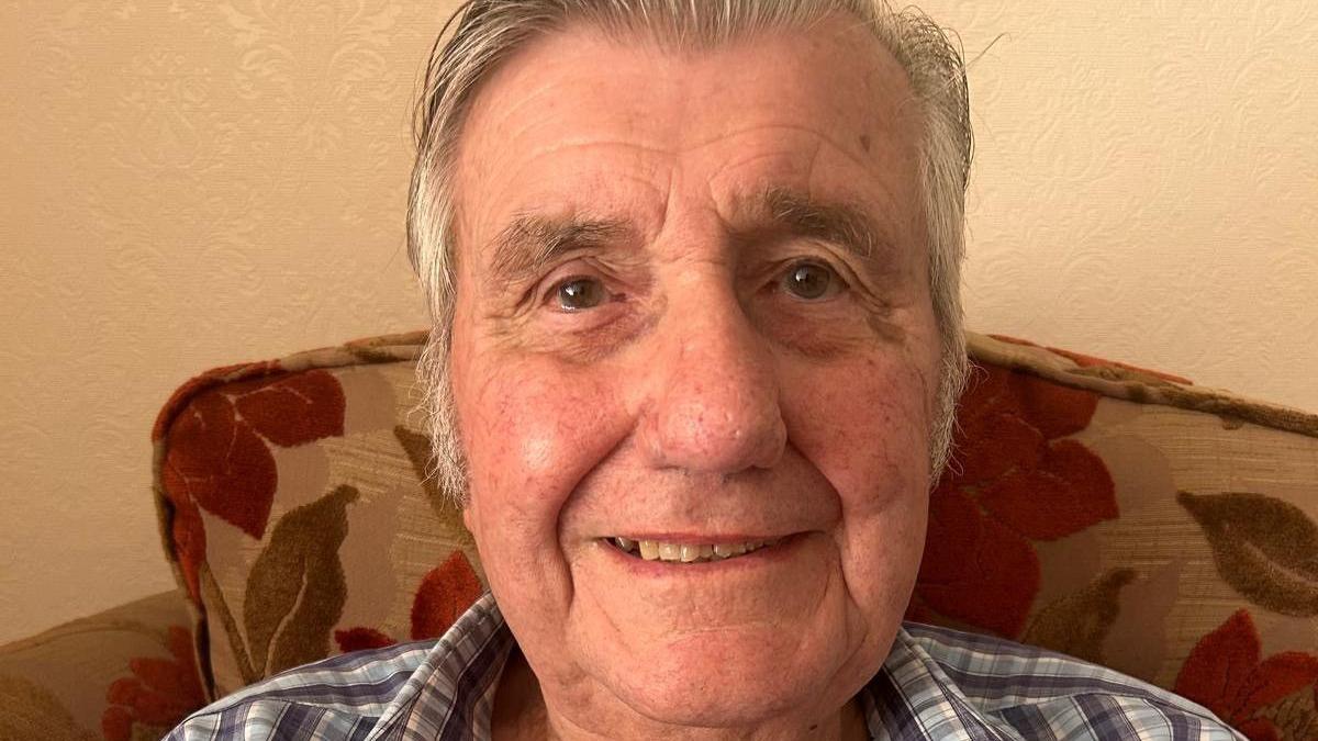 David Judd sits in a living room and smiles at the camera. He has combed grey hair and a check shirt.