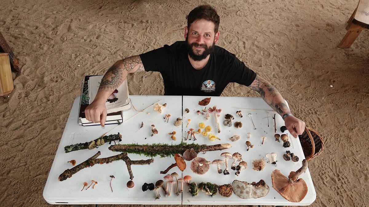 A bird's eye view photo of Andy Knott. He is looking up and smiling for the camera. There is a variety of mushrooms, tree branches and books on a white table in front of him and a basket on the floor beside him. He is wearing a black shirt and has tattoos on his arms. The ground is covered with sand.