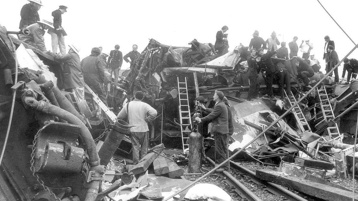 People stand around the wreckage and on top of it. Ladders can be seen leaning against it while pipes and bricks are strewn around.
