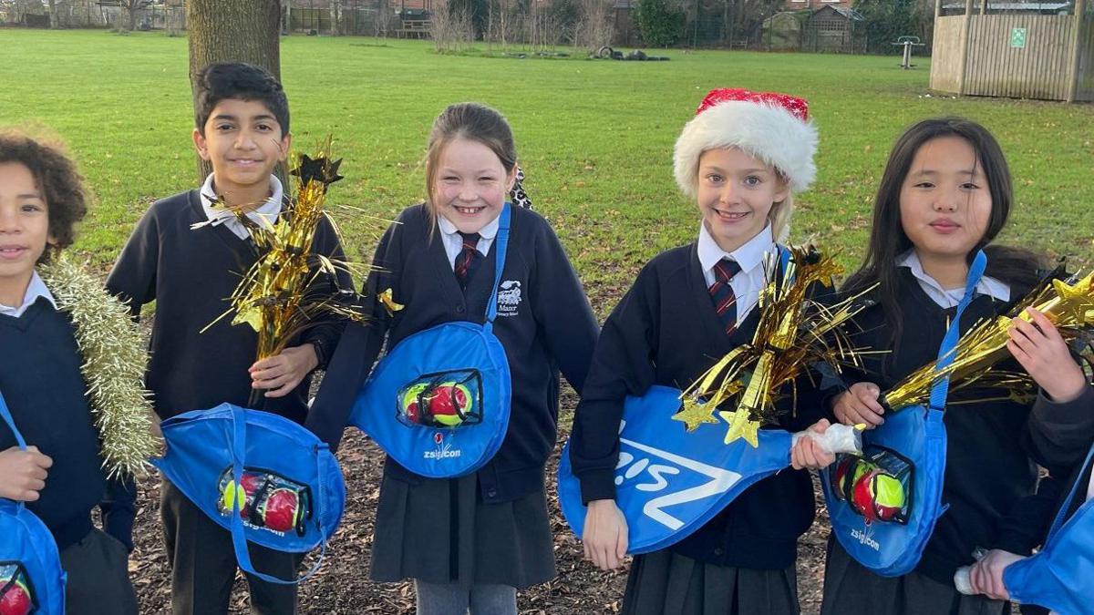 Five schoolchildren, one wearing a Christmas hat, holding tennis rackets in blue bags. They're outside and there's a school field behind them.