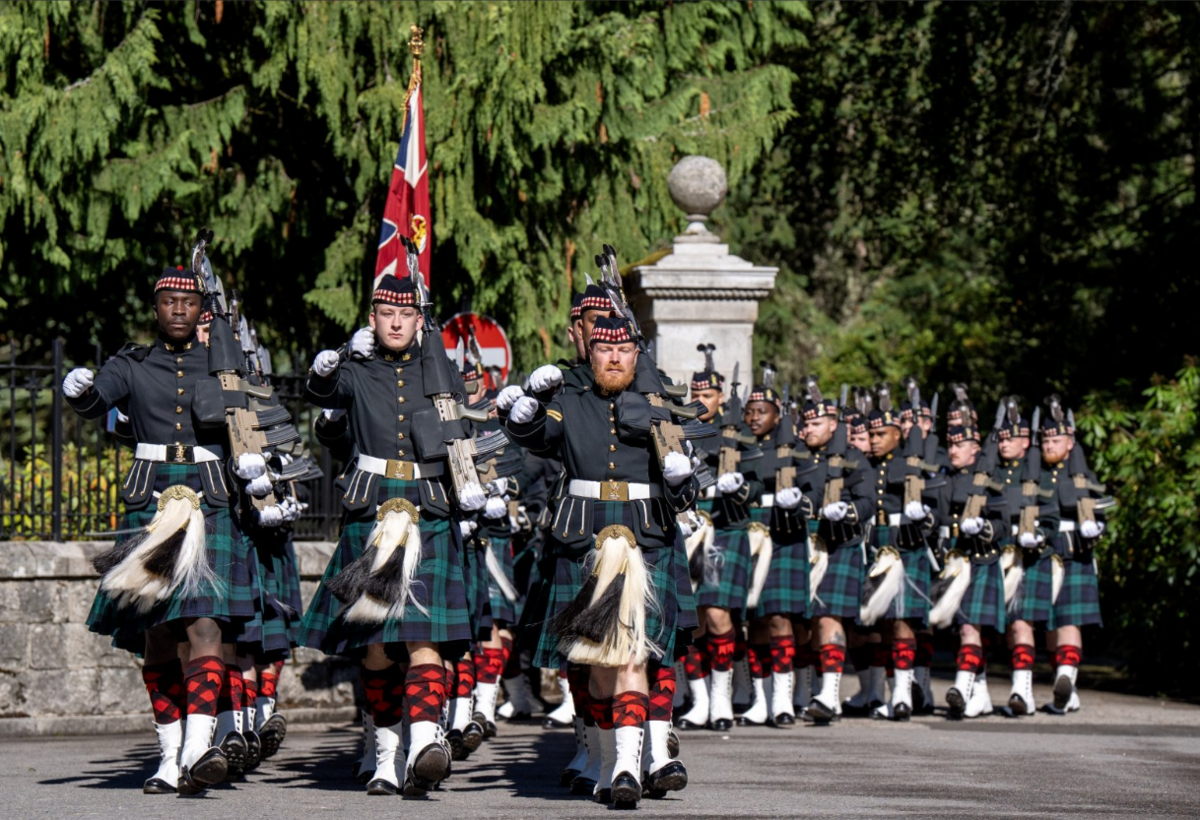 Soldiers from the Royal Regiment of Scotland march in formation during a ceremonial parade, carrying rifles and wearing dark dress uniforms, tartan kilts and white spats. Union flag is carried near the front of the column as the troops advance along a tree-lined route past a stone entrance.