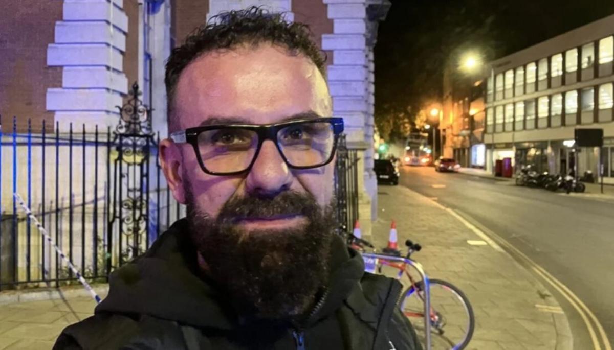 Man with dark curly hair, glasses and a beard looking at the camera. He is stood in a city centre street. It is night time.