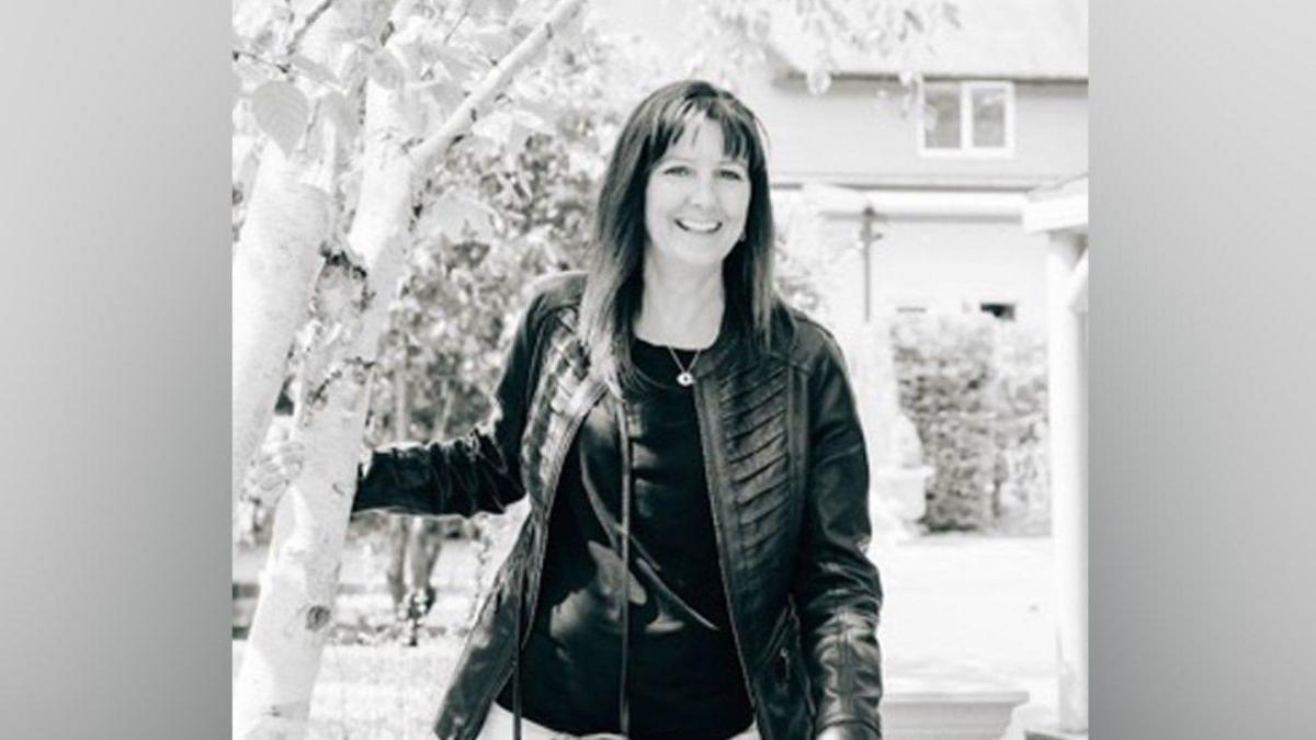A black and white image of a woman with long brown hair and a fringe. She is wearing a leather jacket, black top and a necklace, and is smiling at the camera.
