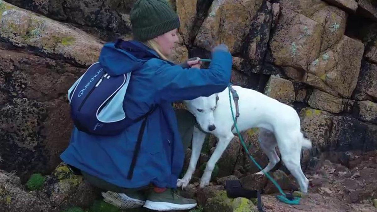 Gail is wearing a blue coat and putting a rope around a white dog on the cliffside