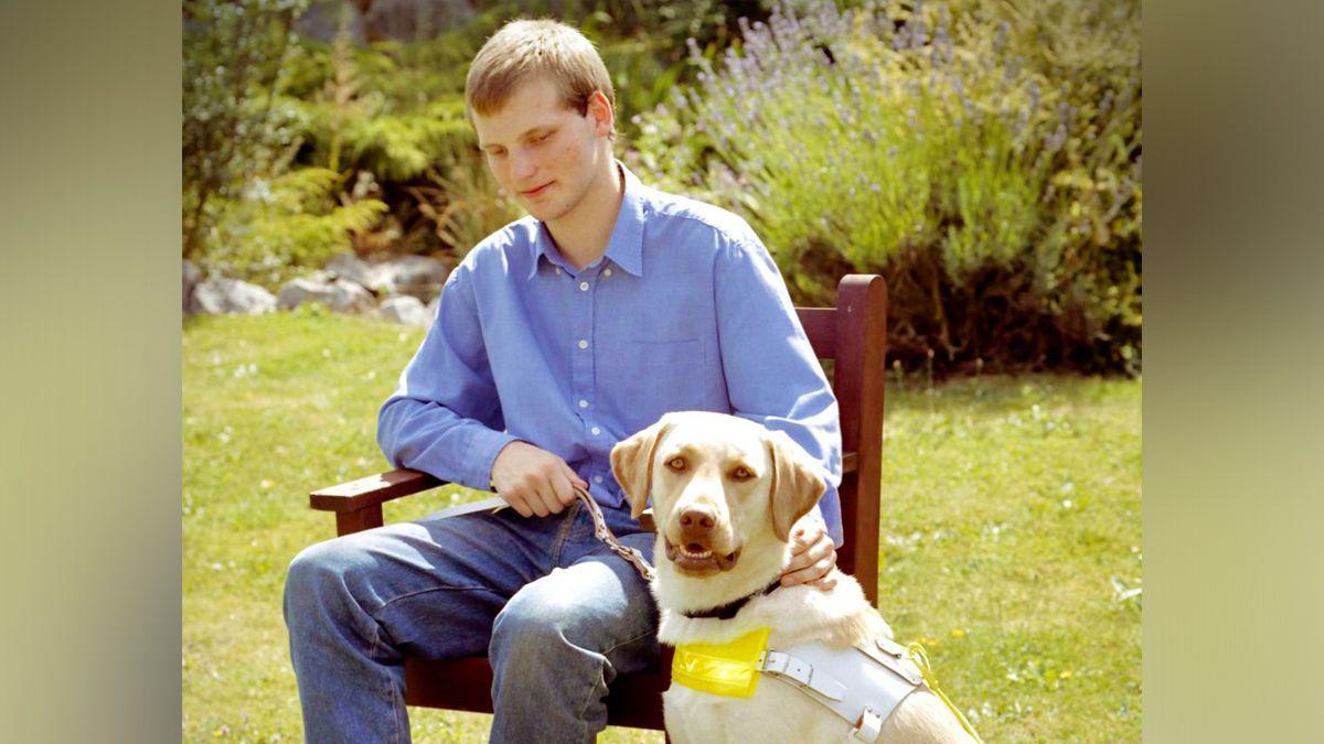 Sean Dilley as a young man, wearing a blue collared shirt and jeans, sitting in a chair in a garden next to his guide dog Brandy