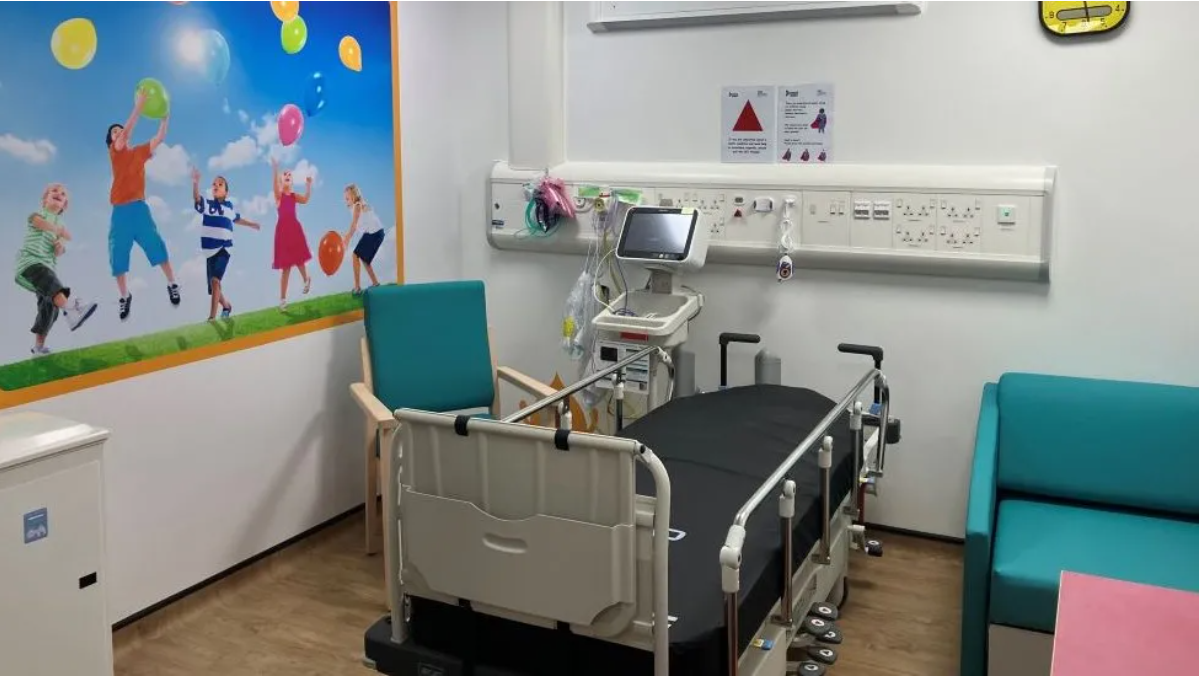 Hospital room with a bed and a blue chair on the left hand side of it while a blue chair is to the right hand side. There is a picture of children playing with balloons on the wall