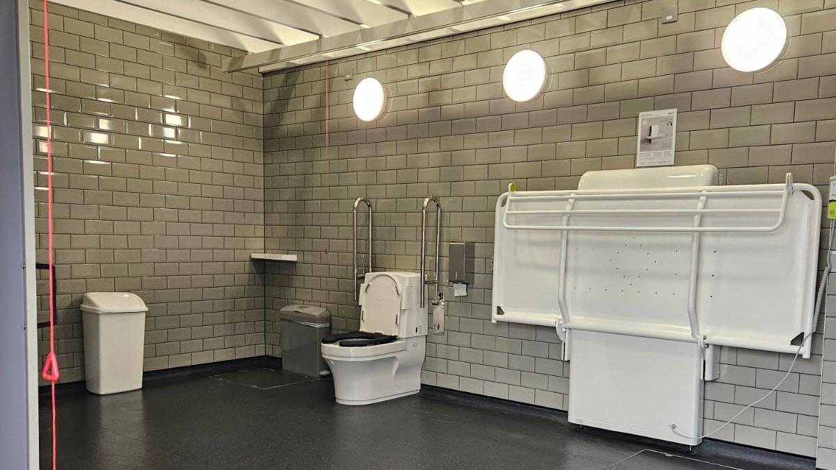 Inside the new changing space, which is a large room with brick shaped sage coloured tiles and a dark grey laminate flooring. There are three circle shaped lights on the right wall of the rectangle shaped room, just above a large pull down table. There is a toilet, bin and red emergency chord against the wall. 