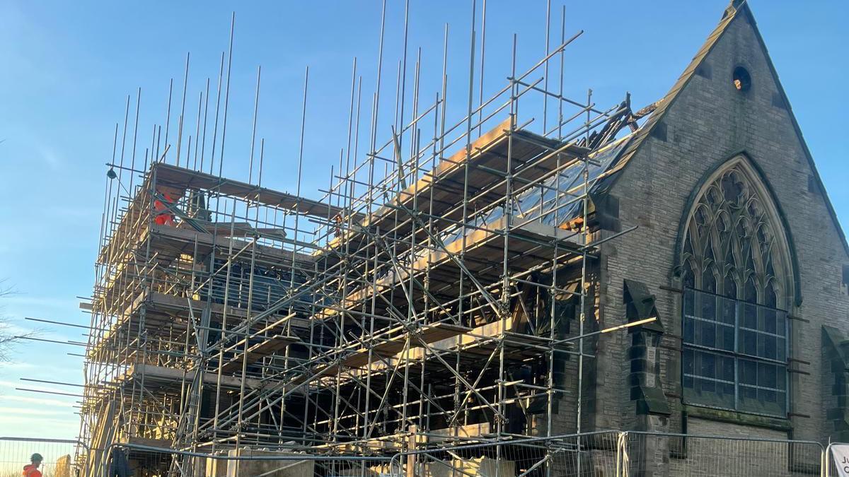 Scaffolding surrounding the outside of the chapel. It has a big stained glass window on the side in the shape of an arch.