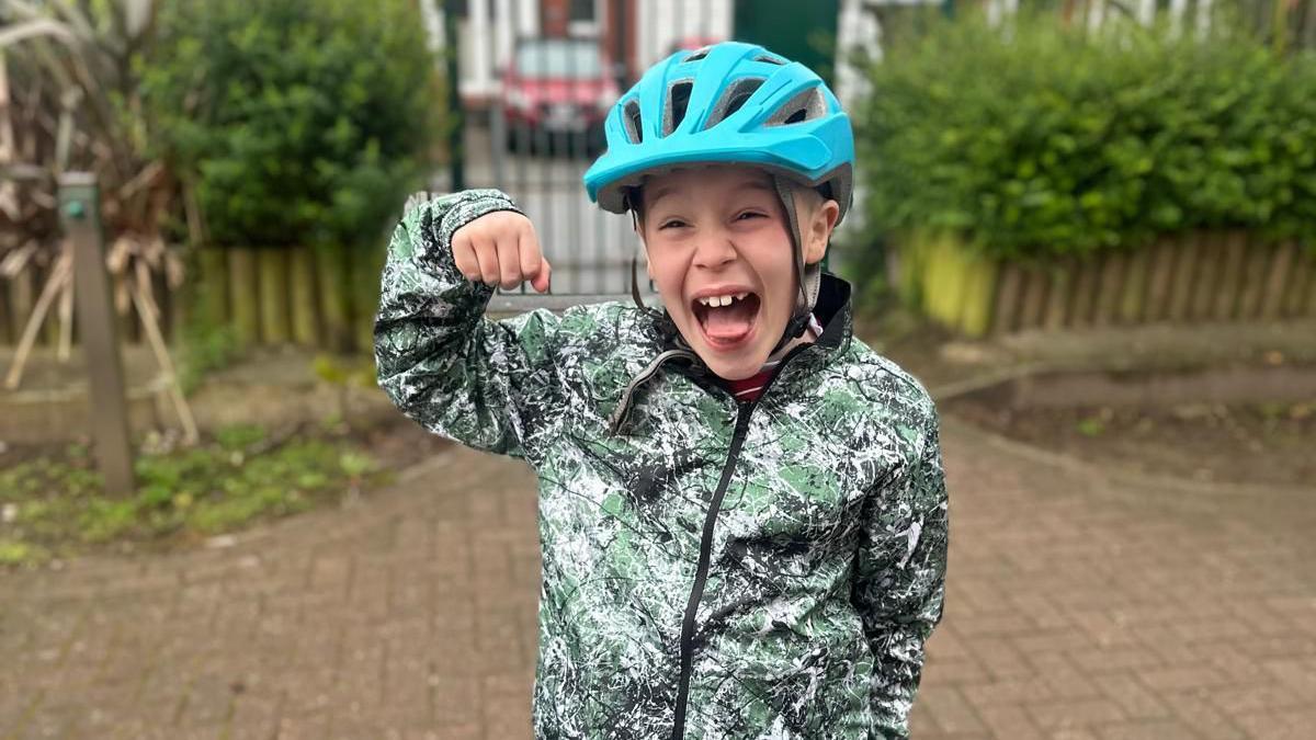 Harvey Goodman, nine, laughs at the camera with his fist raised, while wearing a green and grey patterned coat and a blue bike helmet 