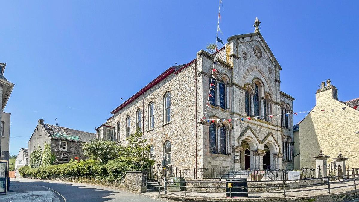 Pre-renovation street view of the former Penryn Methodist Church and School Room where Penryn Art Centre is located. It is a tall stone  church with grey brick and has arched windows at the front and side of the building. There are green trees and bushes to the left of the building and another building behind, and to the right of the church.