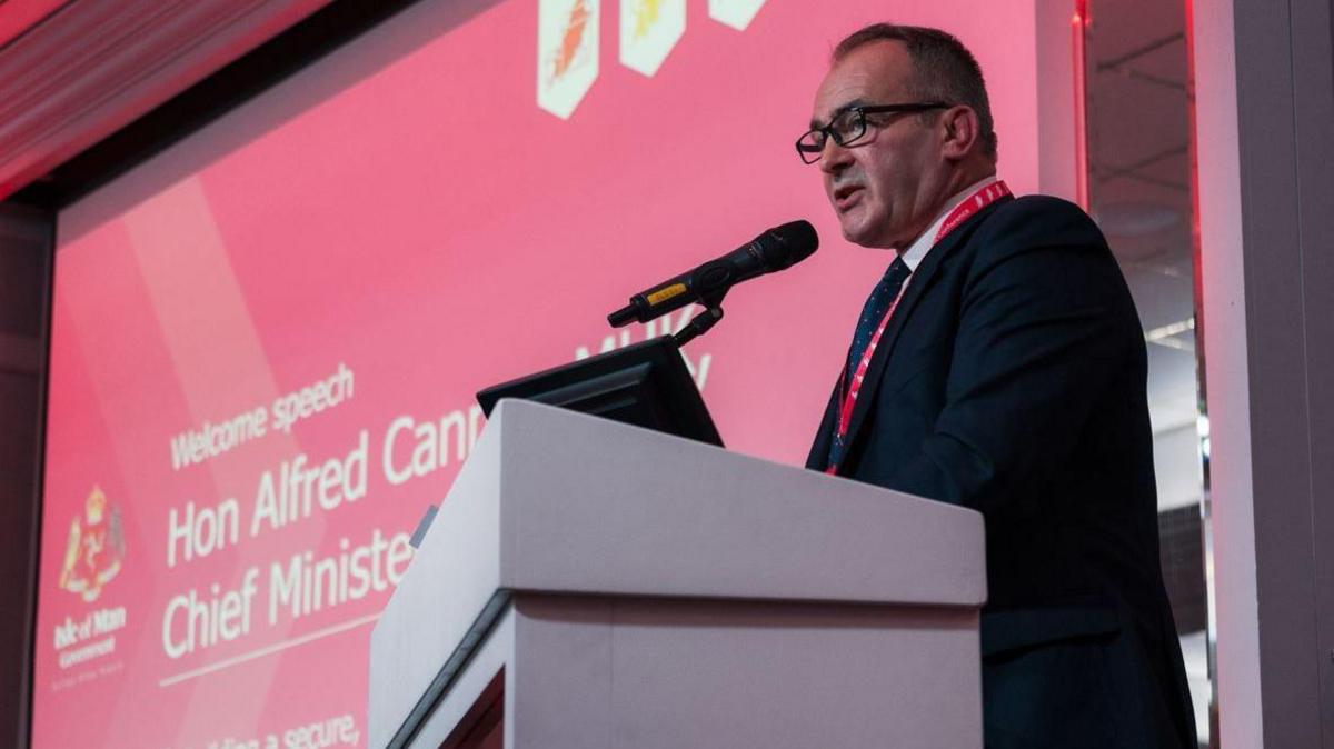 A close-up of the Chief Minister Alfred Cannan as he stands at a lectern delivering a speech speaking into a microphone. He is wearing a dark suit and a large pink screen can be seen behind him.