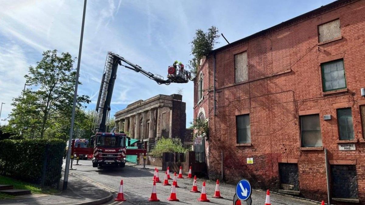 A platform, extended from a fire engine, includes firefighters and is near the building on the right of the photo. Traffic cones are on the road in the foreground.
