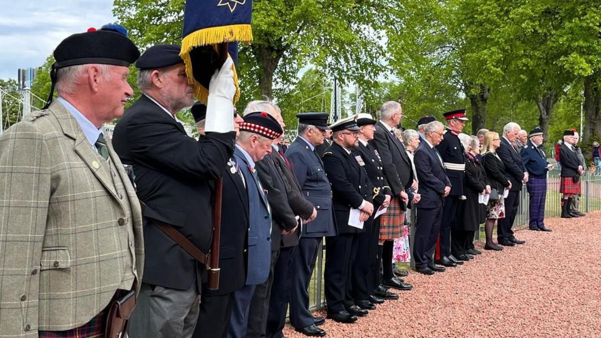 Uniformed veterans stand in a long line of men and a few women with trees and blue sky in the background