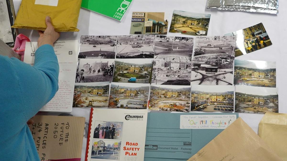 A table covered in documents and A5 photographs. A blue folder in the bottom right reads 'Our 1998 Thoughts'.