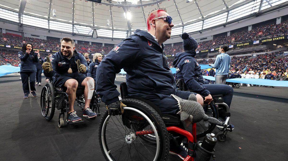 A man in a wheelchair and spiked hair dyed red in the foreground as other teammates in wheelchairs are behind him in the middle of the stadium