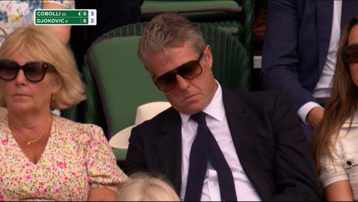 Hugh Grant wears a suit, his tie is skew-whiff and wearing sunglasses. He is asleep