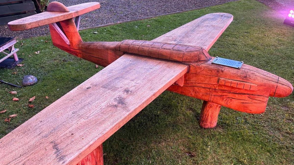 A brown wooden bench carved into the shape of a Fairey Barracuda Torpedo Bomber installed on a patch of grass.