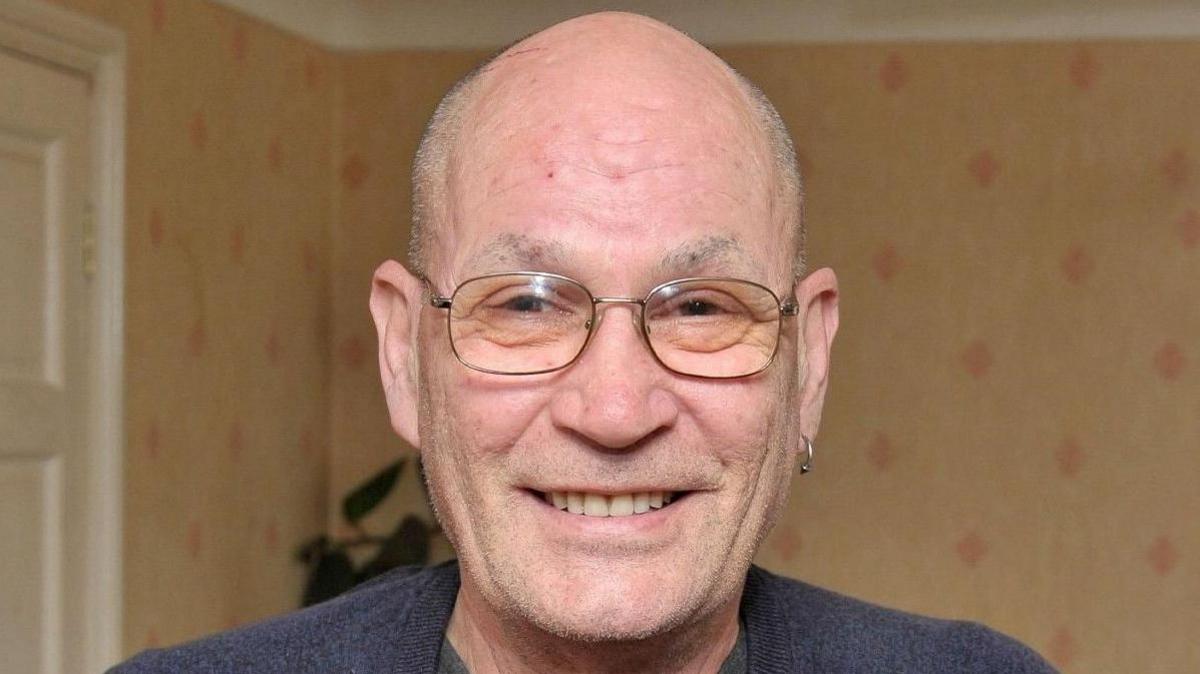 David Newton, wearing a grey sweater over a button-up T-shirt, and metal-framed spectacles, smiles as he holds up a red presentation box embossed with a gold National Blood Service logo and the figure "100" in one hand. In his left ear is an earring. In the other hand is a red soft toy mascot of a blood drop. He is inside what looks like a living room.