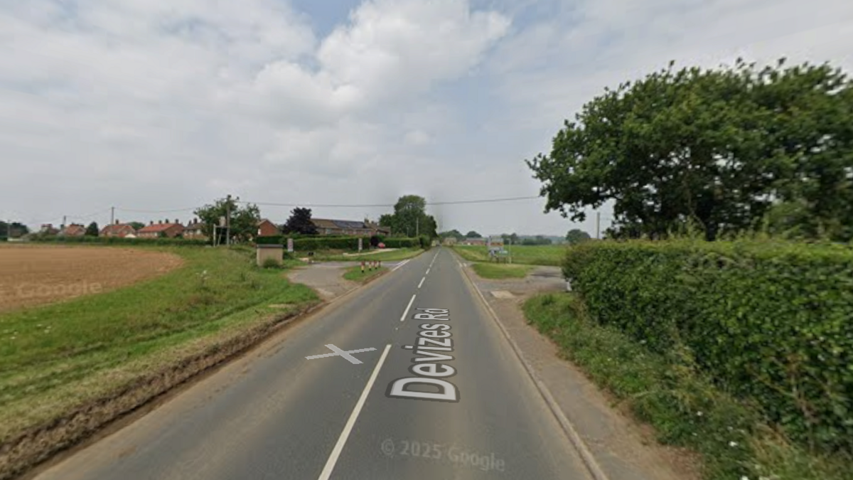 This is a Google Maps image of a road with one lane in each direction. It is a rural scene with a field on one side and a hedge on the other.