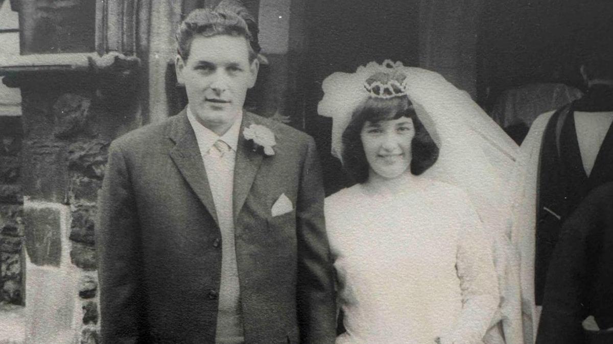 A man wearing a jacket and a woman wearing a white wedding dress standing outside.