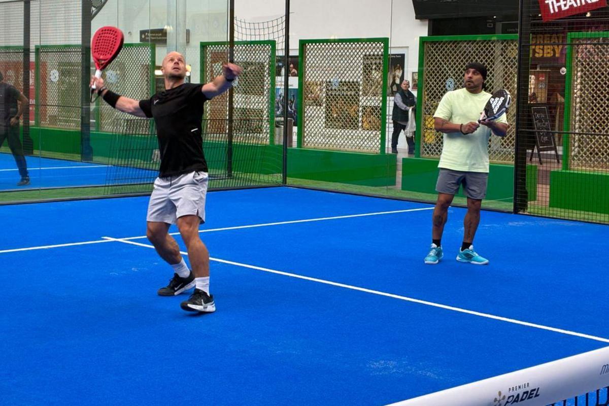 Two people on a blue padel court. One player is looking toward the ceiling about to hit the ball.