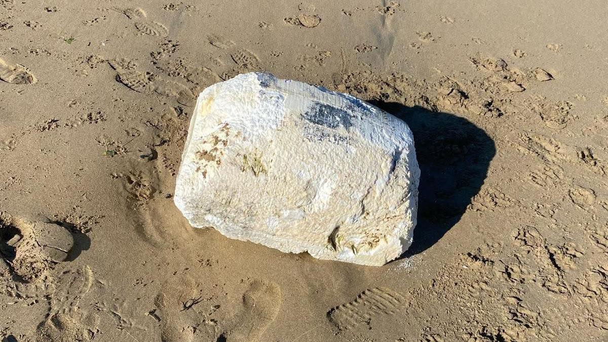 A brick-shaped block of palm oil lying on sand. It is off-white in colour and is surrounded by imprints of feet.