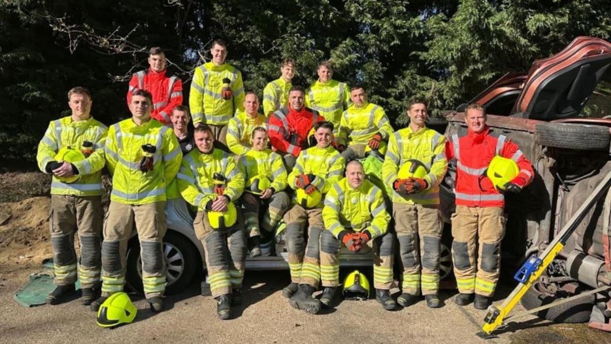 Firefighters from Surrey Fire & Rescue Service pose for a photo. There is a group of 16 people wearing red and orange high-visibility jackets. 