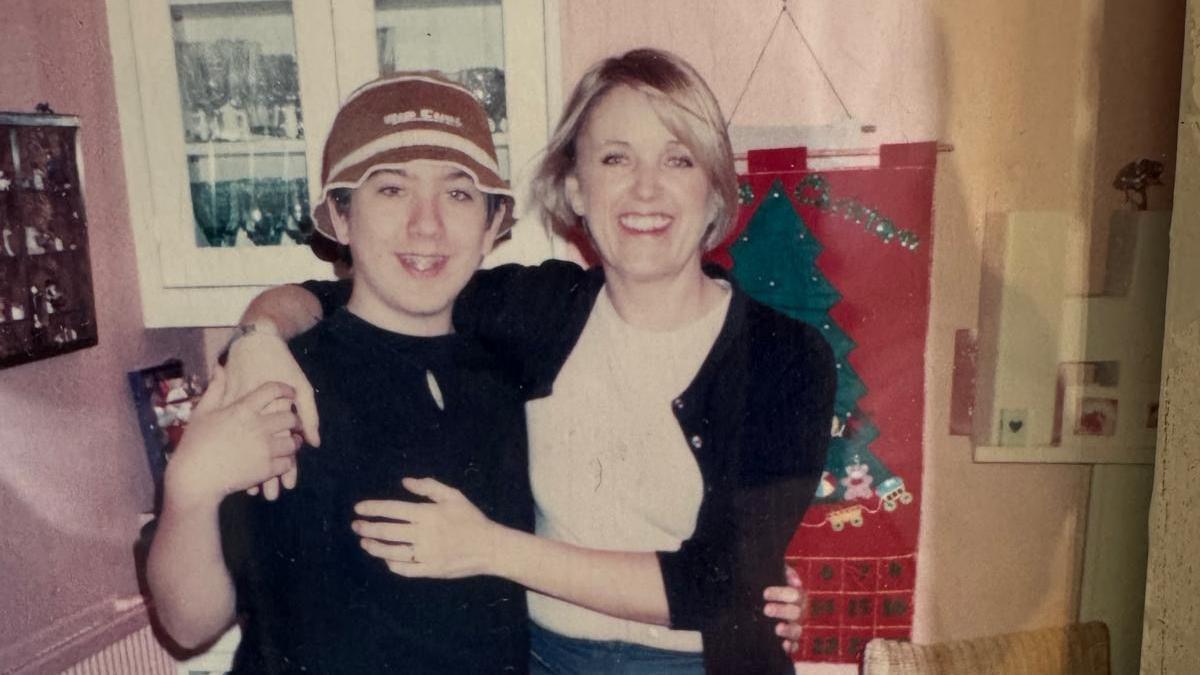 An old photograph showing Poppy hugging her son Alex, they both smile at the camera and alex wears a brown and white hat and a black T shirt. Poppy is in a black cardigan and white top. The photo has been taken in a house and there is a Christmas decoration behind them.