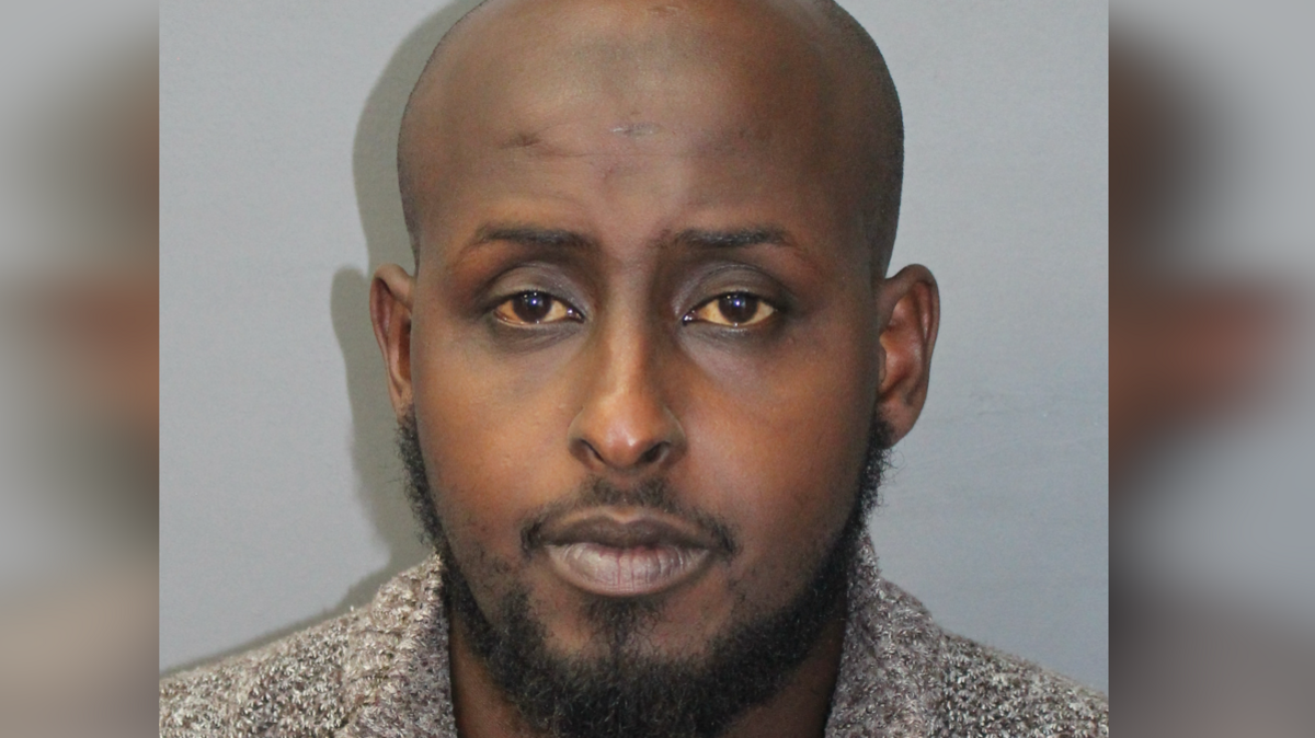 A close up police shot of Mohamed. He is bald with facial hair. Standing against a grey wall. 