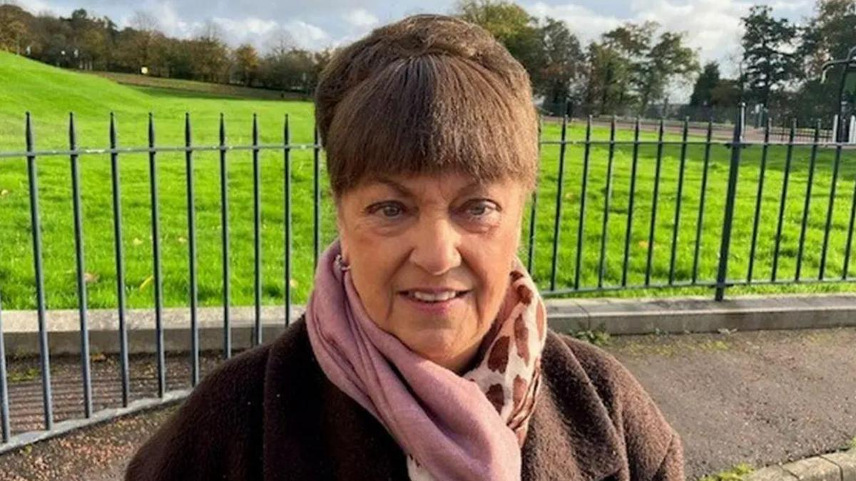 A woman, picture from the shoulders up, looking at the camera. She has brown hair, tied up and has a fringe. She is wearing a brown coat and a scarf. There is a park behind her with steel gates in front of it. 