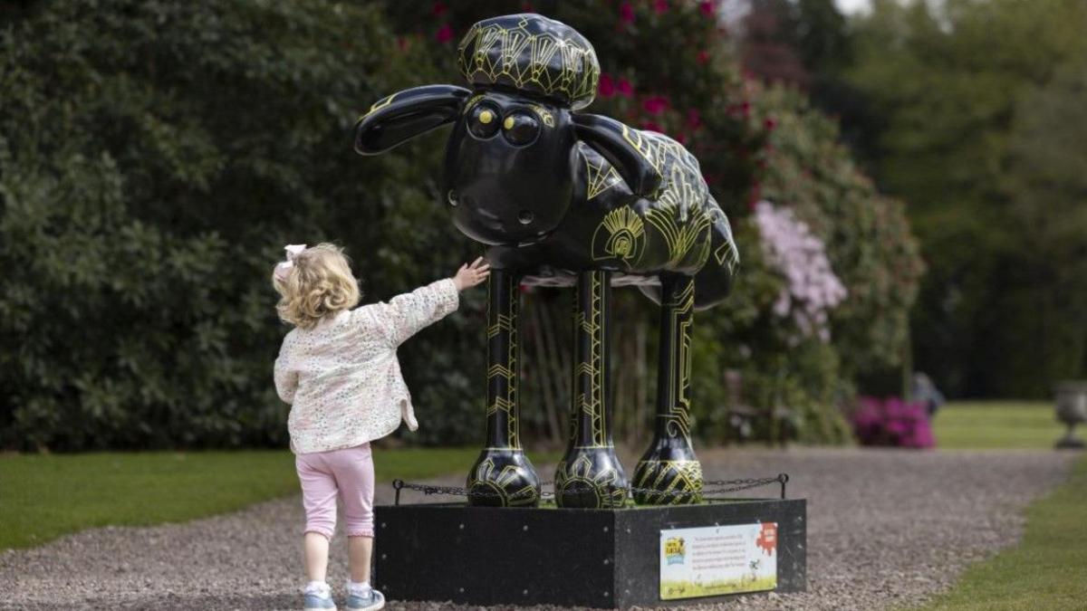 A young girl is reaching up towards a model of a sheep. She has short, curly blonde hair and is wearing a patterned jacket and pink trousers. The sheep is painted black with gold patterns on its body and legs